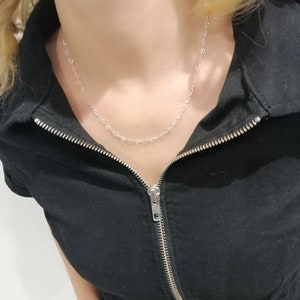 May include: A delicate silver necklace featuring a repeating heart design. The necklace is worn against a black jumpsuit with a silver zipper. The model has blonde hair.