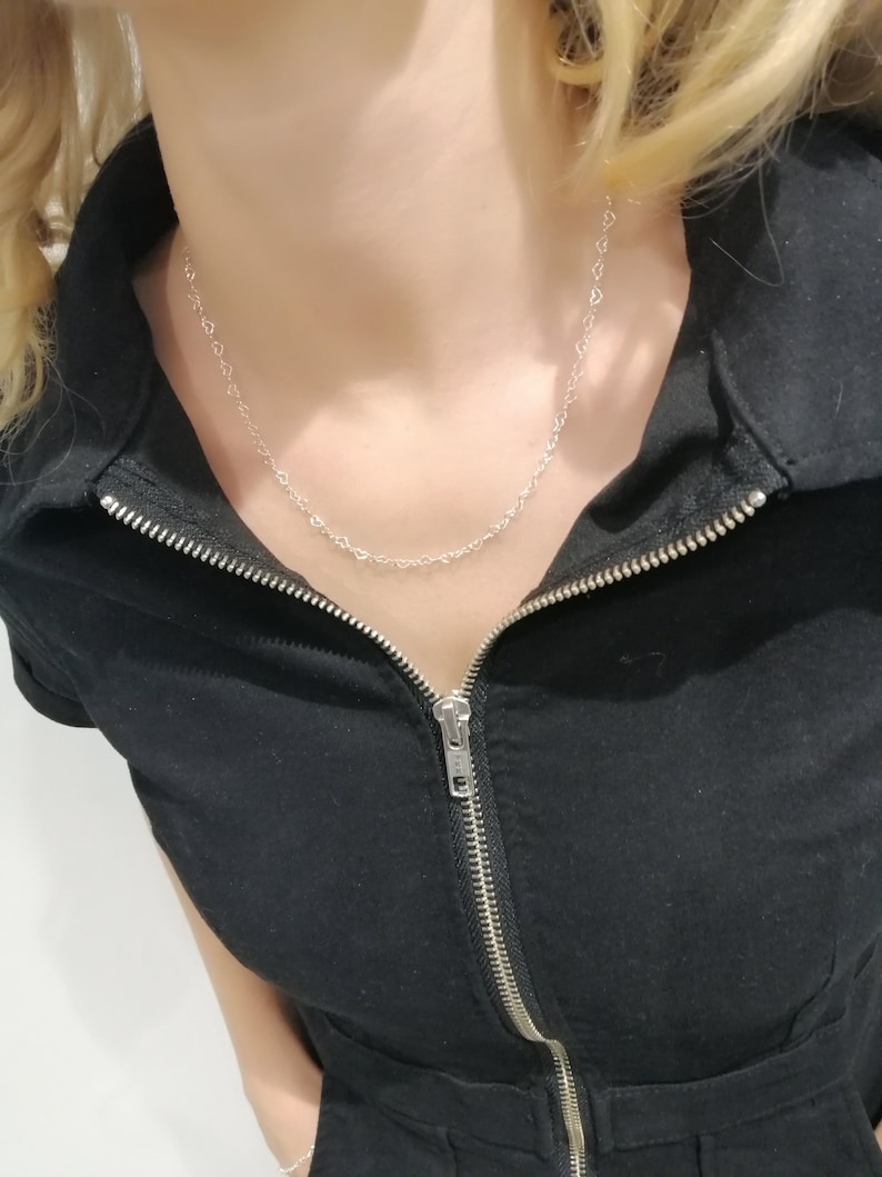 May include: A delicate silver necklace featuring a repeating heart design. The necklace is worn against a black jumpsuit with a silver zipper. The model has blonde hair.