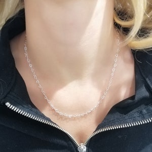 May include: A delicate silver necklace featuring a repeating heart design. The necklace is worn against a black zip-up jacket. The hearts are small and evenly spaced, creating a subtle and elegant accessory.