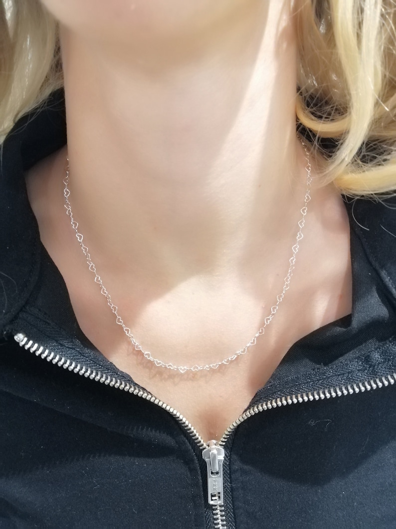 May include: A delicate silver necklace featuring a repeating heart design. The necklace is worn against a black zip-up jacket. The hearts are small and evenly spaced, creating a subtle and elegant accessory.