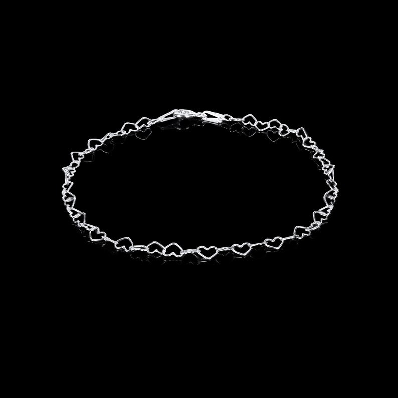May include: A delicate silver bracelet featuring a repeating heart design. The bracelet is composed of small, open-heart shapes linked together, creating a romantic and elegant accessory. The clasp is visible at the top of the bracelet.