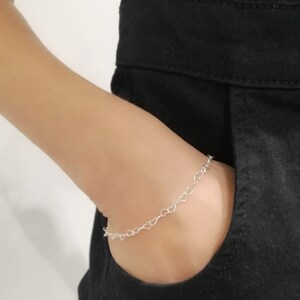 May include: A delicate silver bracelet featuring a series of small heart-shaped links. The bracelet is worn on a wrist, with a black garment in the background. The bracelet is a simple yet elegant piece of jewelry.