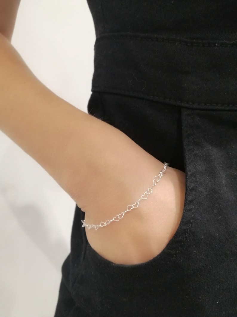 May include: A delicate silver bracelet featuring a series of small heart-shaped links. The bracelet is worn on a wrist, with a black garment in the background. The bracelet is a simple yet elegant piece of jewelry.