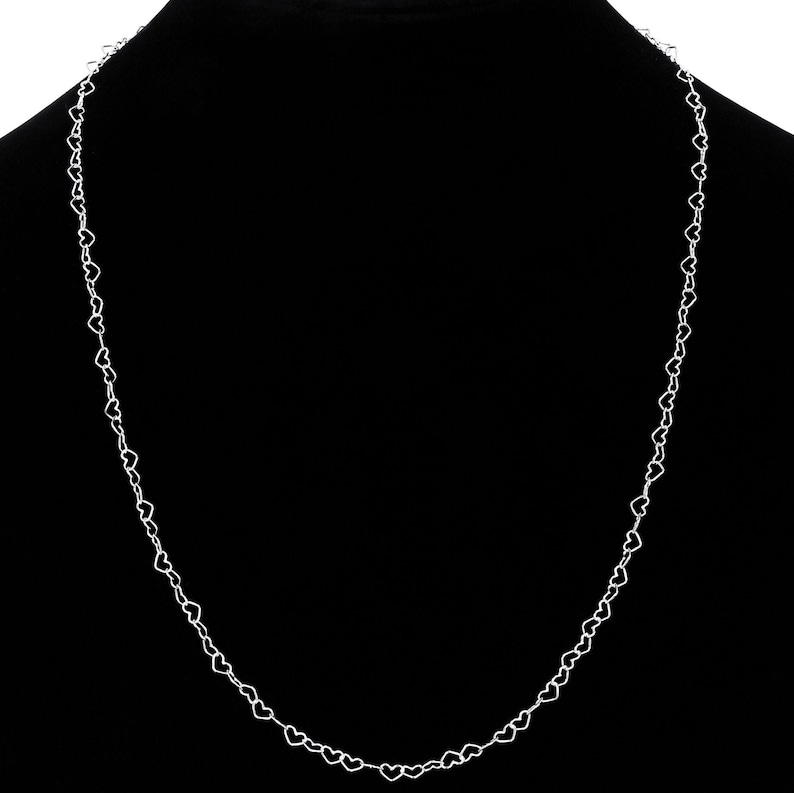 May include: A delicate silver necklace featuring a repeating heart design. The necklace is displayed against a black background, highlighting the intricate details of the heart-shaped links. The chain is lightweight and appears to be made of a shiny metal.