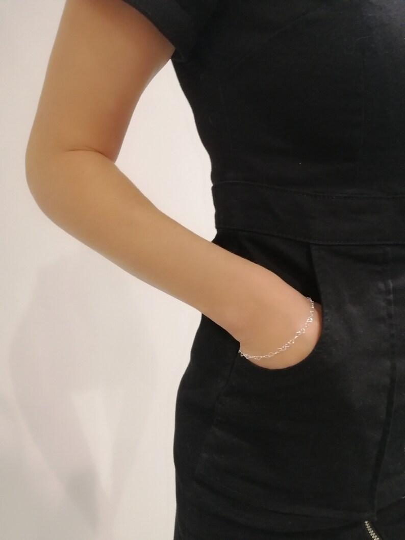 May include: A delicate silver bracelet with a heart-shaped design is visible on the wrist. The bracelet is worn with a black jumpsuit. The person's arm is in the pocket of the jumpsuit.