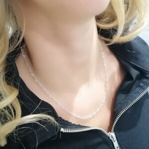 May include: A delicate silver necklace featuring a repeating heart design. The necklace is worn against a black jacket, creating a contrast that highlights the jewelry. The chain is fine and the hearts are small, creating a subtle and elegant look.