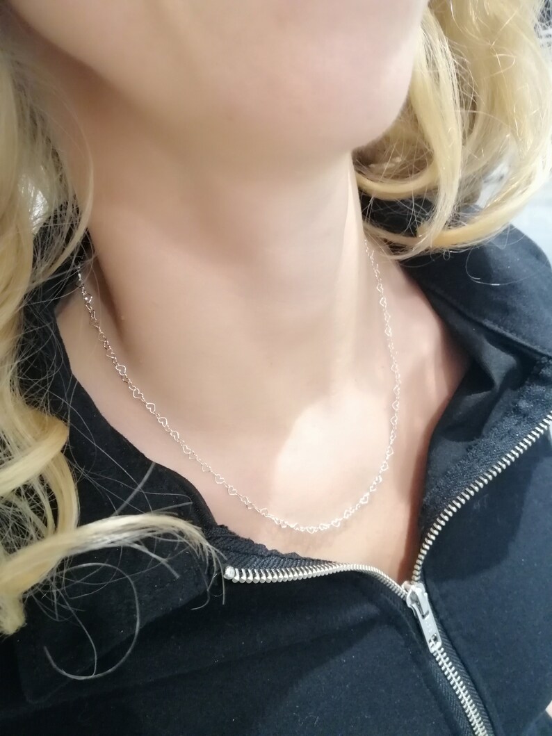 May include: A delicate silver necklace featuring a repeating heart design. The necklace is worn against a black jacket, creating a contrast that highlights the jewelry. The chain is fine and the hearts are small, creating a subtle and elegant look.