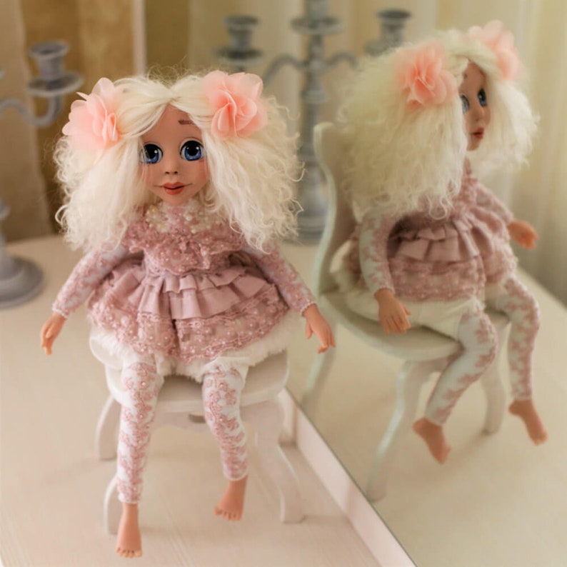 May include: A handmade doll with white curly hair, wearing a pink dress with lace details and white leggings. The doll is sitting on a white chair with a pink flower in her hair.