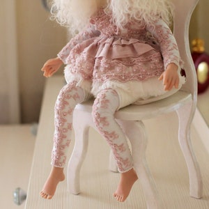 May include: A porcelain doll wearing a pink and white lace dress and white leggings with pink floral embroidery. The doll is sitting on a white wooden chair.