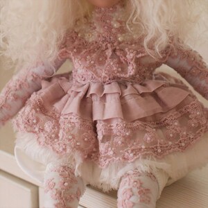 May include: A pink doll wearing a lacy dress with pearl embellishments. The dress has a ruffled skirt and is layered with tulle. The doll has long, white, curly hair.