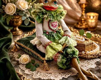 May include: A whimsical frog doll with a green and white ruffled collar and dress, seated on a green velvet pillow. The doll has a red mouth and is adorned with beaded accents. A decorative gold tray and a bouquet of white roses are in the background.