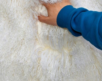 May include: White fluffy sheepskin rug with long, soft fibers. The rug is being touched by a hand wearing a blue sleeve.