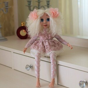 May include: A handmade doll with white curly hair, wearing a pink lace dress and leggings. The doll is sitting on a white dresser with drawers.