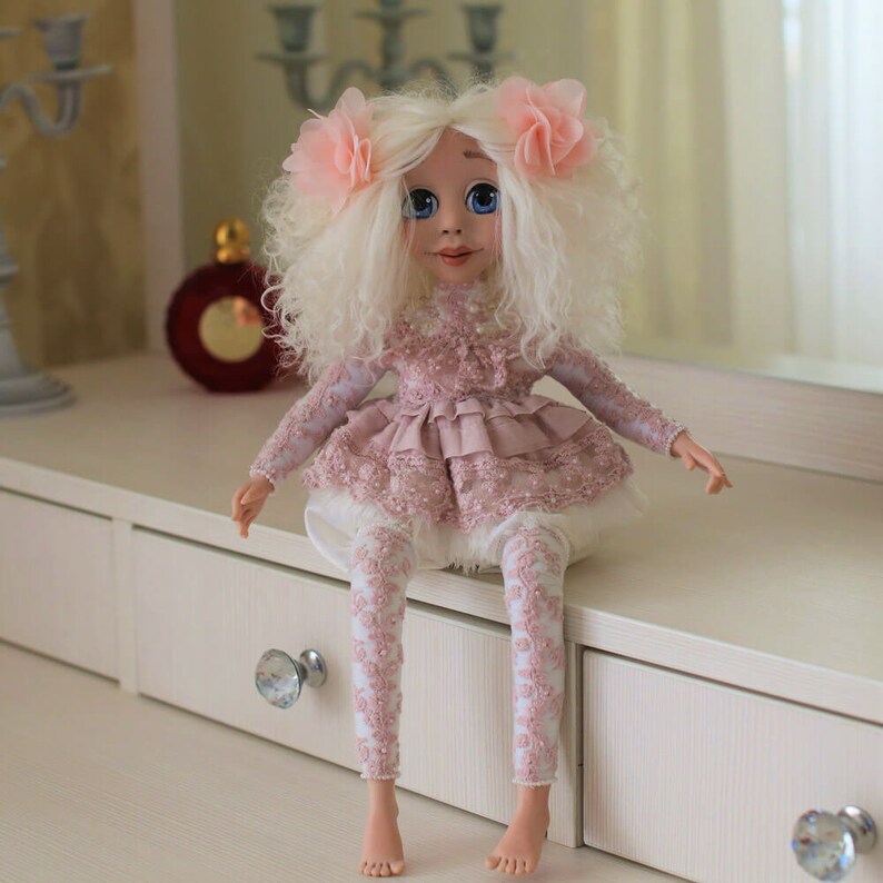 May include: A handmade doll with white curly hair, wearing a pink lace dress and leggings. The doll is sitting on a white dresser with drawers.