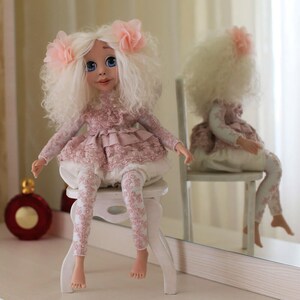 May include: A white doll with curly hair and pink bows sits on a white stool wearing a pink dress and leggings with lace details.