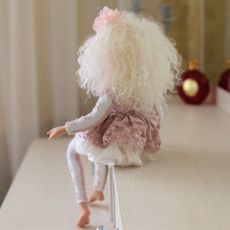 May include: A porcelain doll with long, white curly hair wearing a pink and white dress with lace details. The doll is sitting on a white surface.