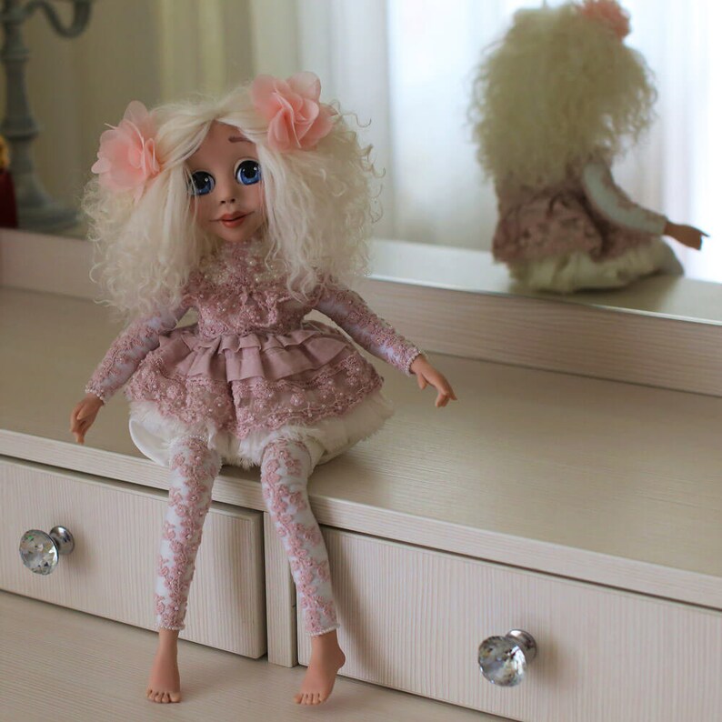 May include: A handmade doll with white curly hair, wearing a pink dress with lace and white leggings. The doll is sitting on a white dresser with two drawers.