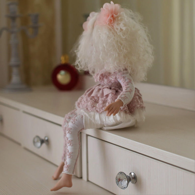May include: A doll with white curly hair wearing a pink dress with lace trim and white tights. The doll is sitting on a white dresser with drawers.