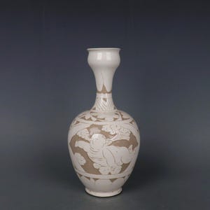 Könnte beinhalten: Eine weiße Keramikvase mit langem Hals und bauchigem Körper. Die Vase hat ein detailliertes Reliefdesign einer Figur und floralen Mustern in einer kontrastierenden beigen Farbe. Die Vase hat eine glatte, glänzende Oberfläche.