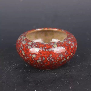 Könnte beinhalten: Ein kleiner, runder Keramik-Pflanztopf mit glänzender, roter Glasur, die mit grünen und braunen Flecken versehen ist. Der Innenraum ist hellbraun. Der Pflanztopf hat eine breite Öffnung und ein niedriges Profil, geeignet für kleine Pflanzen oder Sukkulenten.