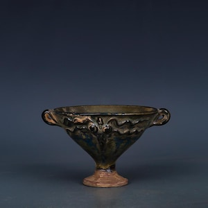 May include: A dark-colored, antique-style ceramic bowl with a flared, pedestal base and two small handles. The bowl has a textured surface with a mix of green, blue, and brown hues, suggesting an aged appearance. The base is a lighter brown.