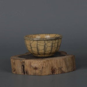 Może przedstawiać: Mała, kremowa ceramiczna miska z pękniętym szkliwem. Miska stoi na drewnianym stojaku.