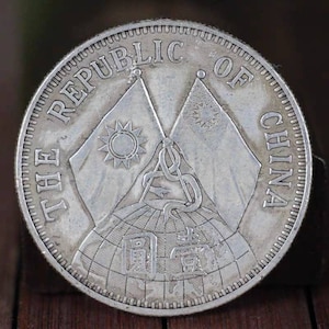 以下が含まれることがあります： 縁に「THE REPUBLIC OF CHINA」の文字が入った銀貨。太陽のシンボルが付いた2つの旗が、中国語の文字が書かれた地球儀の上で交差しています。コインは詳細で質感のあるデザインです。