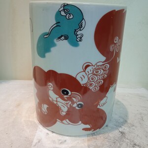 May include: White ceramic cylinder vase with a red and blue design. The design features two stylized animals, one red and one blue, with swirling patterns.