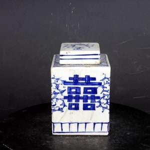 May include: A white porcelain square tea caddy with a blue and white floral design and the Chinese character for "double happiness" on the front. The lid is also decorated with blue and white floral designs.
