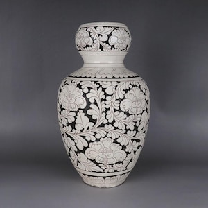 Könnte beinhalten: Eine hohe, weiße Keramikvase mit einem bauchigen Körper und einem kleineren, abgerundeten oberen Teil. Die Vase ist mit einem detaillierten schwarzen Blumenmuster verziert, das Blumen und Blätter umfasst und einen auffälligen Kontrast erzeugt.
