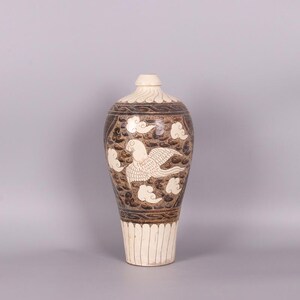 May include: A tall, decorative ceramic vase with a cream-colored base and top. The vase features a brown body with a white bird and cloud design. The bird is in flight, and the clouds are stylized.