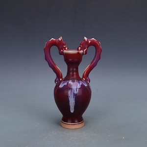 May include: A maroon ceramic vase with two dragon-shaped handles. The handles are a gradient of purple and red. The vase has a white and purple design on the front and a beige base.