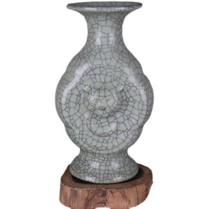 Puede incluir: Un jarrón de cerámica verde claro con un esmalte craquelado y una forma redondeada única. El jarrón está decorado con un diseño en relieve y se asienta sobre una base de madera.