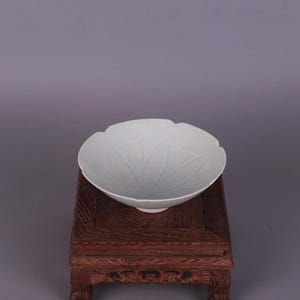 May include: A white porcelain bowl with a scalloped edge and a floral design. The bowl is placed on a small, dark wooden stand. The bowl's design features a delicate, embossed leaf pattern.