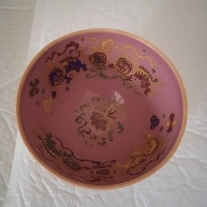 Puede incluir: Un cuenco de cerámica rosa con un borde dorado y diseños intrincados en oro de criaturas míticas y motivos florales. El cuenco tiene un interior rosa claro.