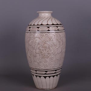 May include: A tall, white ceramic vase with a detailed, hand-painted design. The vase features a repeating floral pattern in brown, with decorative bands at the top, middle, and base. The vase has a narrow neck and a rounded body.