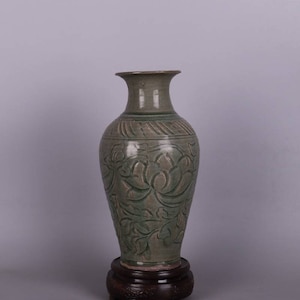 May include: A tall, green ceramic vase with a carved floral design. The vase has a narrow neck and a wide base. It is sitting on a wooden stand.