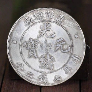 以下が含まれることがあります： 表に「光緒元寶」 (Guangxu Yuanbao) と「大清銀幣」 (Da Qing Yinbi) の文字が刻まれた銀製の中国の硬貨。硬貨はビーズの縁取りで囲まれています。
