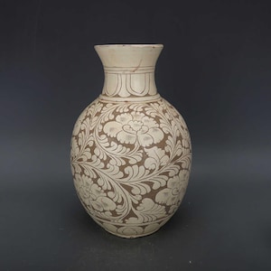 Könnte beinhalten: Eine cremefarbene Vase mit einem braunen Blumenmuster. Die Vase hat einen breiten, abgerundeten Körper und einen schmalen Hals. Das florale Design bedeckt den gesamten Körper der Vase.