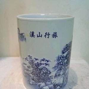 Puede incluir: Un recipiente cilíndrico de cerámica con fondo azul claro y diseño de paisaje azul oscuro. El diseño incluye árboles, rocas y figuras, con caracteres chinos. Probablemente un objeto decorativo o un jarrón.