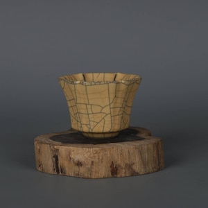 Könnte beinhalten: Eine hellgelbe Keramiktasse mit craquelé-Glasur und einem gewellten Rand. Die Tasse steht auf einem Holzsockel.
