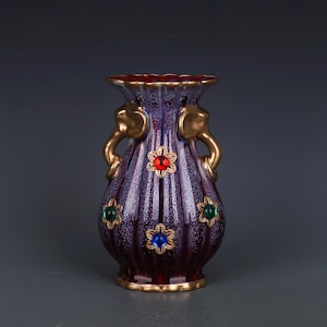 May include: A decorative vase with a deep purple and burgundy color scheme. The vase features gold-colored elephant head handles and is adorned with red, green, and blue jewel-like accents. The rim and base are trimmed with gold.