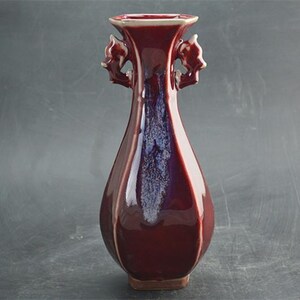May include: A tall, burgundy-colored ceramic vase with a flared top and two ornate handles. The vase has a glossy finish with a subtle blue and purple iridescence. The base is a darker brown color.