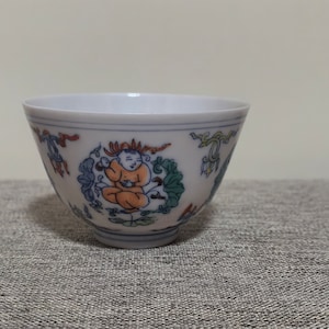 Puede incluir: Una taza de té de cerámica blanca con un diseño floral azul y verde y una pequeña figura naranja en el centro.