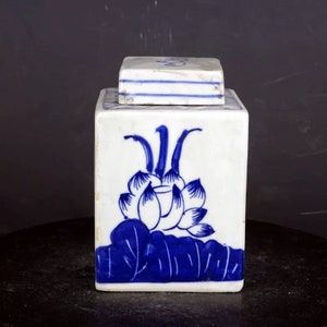 May include: A white ceramic square jar with a blue and white lotus flower design. The jar has a lid and is decorated with blue lines.