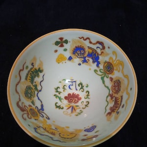 Puede incluir: Un cuenco de cerámica blanca con un borde amarillo y diseños intrincados pintados a mano. El cuenco presenta un motivo central de una nube roja con un patrón floral azul y verde, rodeado de cuatro leones estilizados en azul, amarillo y rojo.