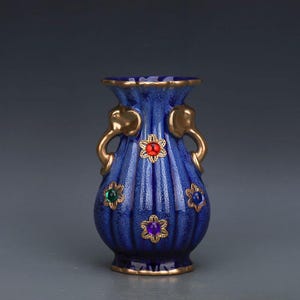 May include: A blue ceramic vase with gold accents and decorative gold handles. The vase features vertical ridges and is adorned with colorful floral-shaped jewels. The rim and base are trimmed with gold.