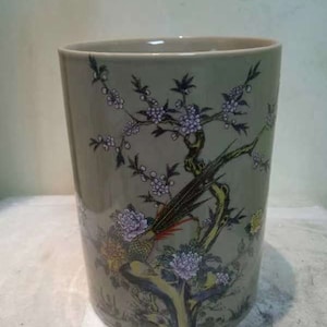 Puede incluir: Un jarrón de cerámica gris con un diseño floral que presenta un pájaro y ramas con flores moradas. El jarrón tiene una boca ancha y un fondo redondeado.