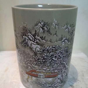 Puede incluir: Un jarrón de cerámica gris con una escena de invierno pintada en él. La escena presenta un paisaje nevado con árboles, una casa y personas.