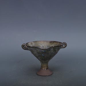 May include: A handmade ceramic bowl with a flared rim and two small handles. The bowl has a dark, mottled glaze with hints of blue and green. It sits on a pedestal base with a brown finish. The bowl's design is rustic and artistic.